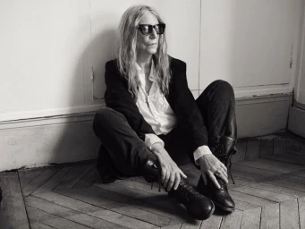 Patti Smith wearing dark eyewear, a black jacket, white shirt, and boots while seated on a chevron wood floor.