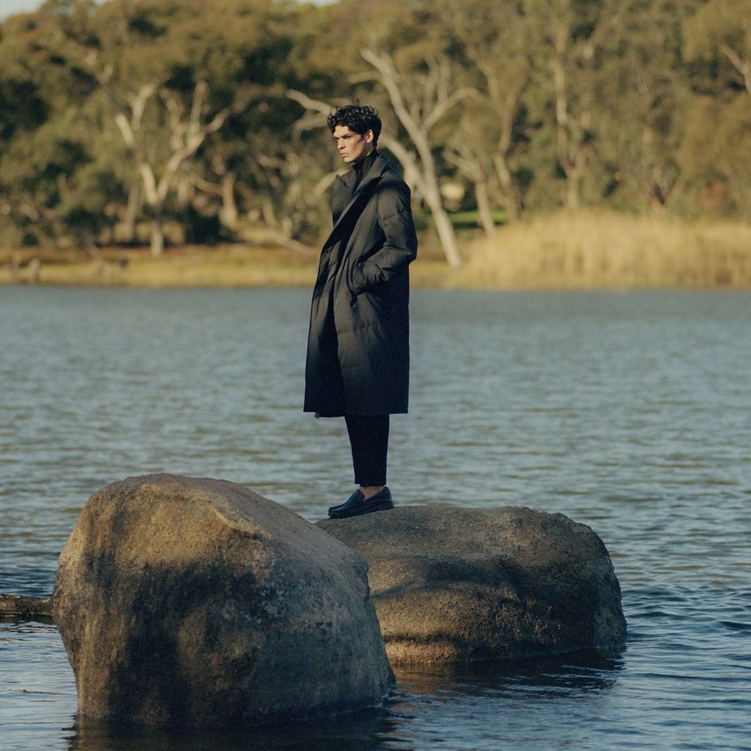 Man standing on a large rock in the water wearing a dark coat and trousers with trees in the background