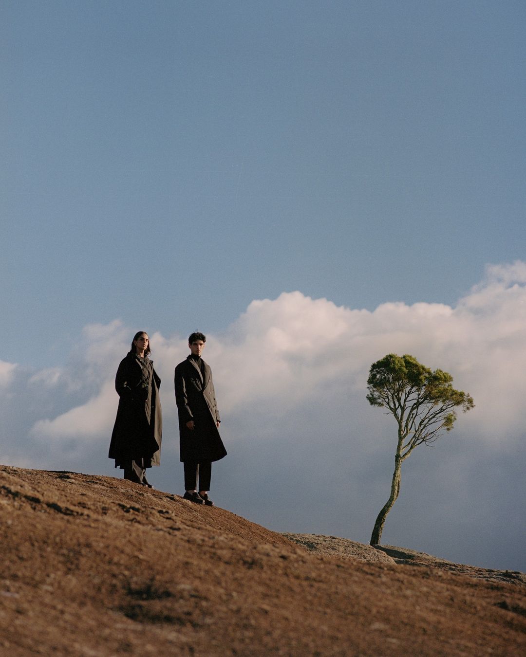 Two models wearing dark coats standing on a hillside with a small tree under an open sky