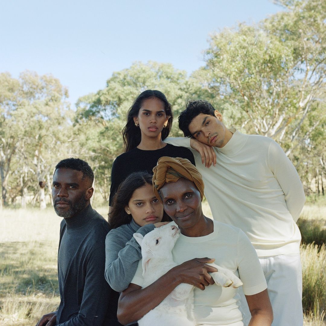 Five people pose in an outdoor grassy setting with trees. The woman in the foreground holds a white lamb. Medium shot.
