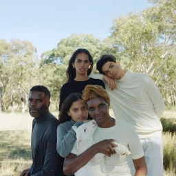Group portrait of five people posing outdoors in a grassy field with trees, featuring an older woman holding a white lamb.