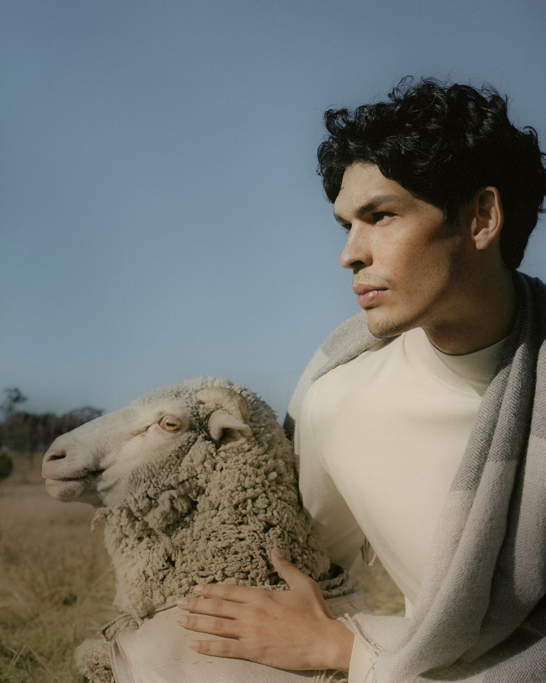 Man with dark curly hair wearing a cream top and wool scarf standing outdoors next to a sheep under a clear blue sky