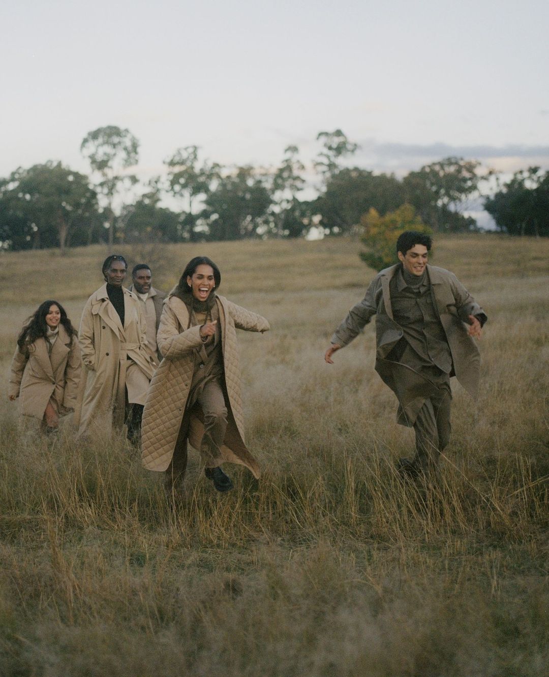 A group of five people running through a grassy field. The people are wearing tan and brown clothing, and trees are in the background.