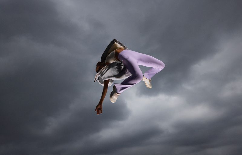 A woman in a white tank top and lavender pants is mid-air against a stormy sky, wearing white sneakers and a grey jacket.