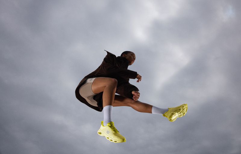 Athletic person mid-air jumping with legs splayed out wearing brown jacket, beige shorts, white socks, and yellow sneakers against cloudy sky.