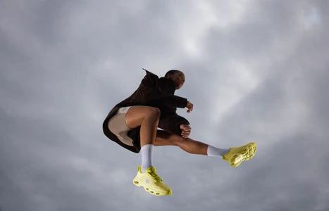 Low-angle shot of a person leaping mid-air against an overcast sky, wearing bright yellow sneakers, white socks, and a dark jacket.