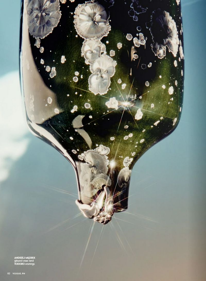 Close-up view of a glossy dark green glass object with scattered white spots, with highlights and a droplet against a blue gradient background.