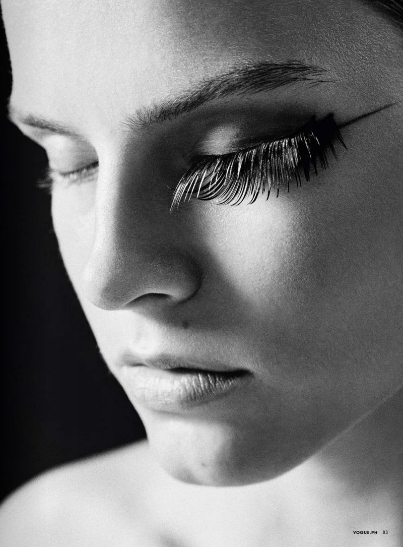 Close-up portrait of a woman with striking false eyelashes, black eyeliner, closed eyes, and pale skin against a dark background.