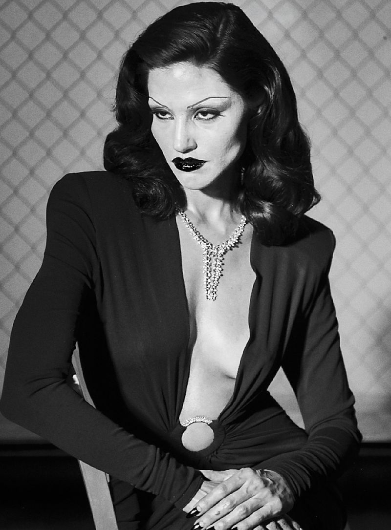 Monochrome portrait of a woman with bold makeup wearing a long-sleeved dress and necklace sitting indoors with patterned background.