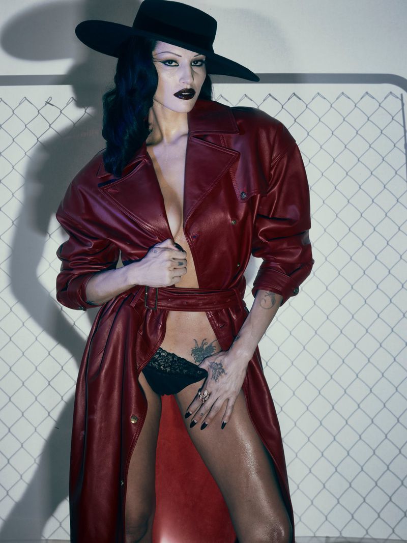 Striking fashion shot of a woman in a red leather coat and black hat against a patterned backdrop, showcasing bold makeup and a confident pose.