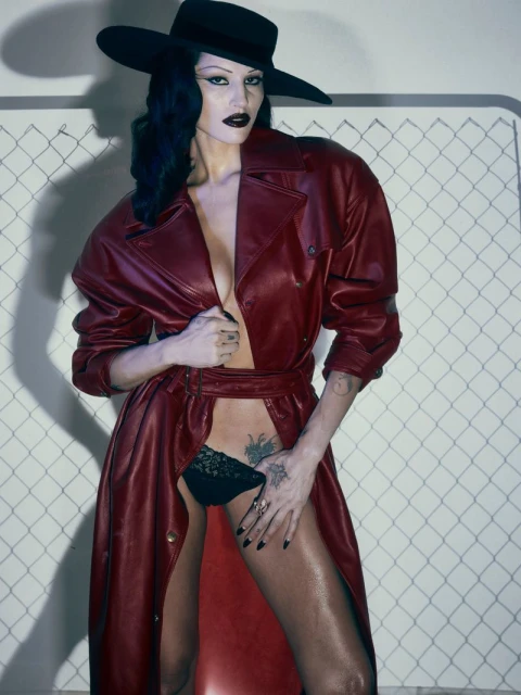 Striking fashion shot of a woman in a red leather coat and black hat against a patterned backdrop, showcasing bold makeup and a confident pose.