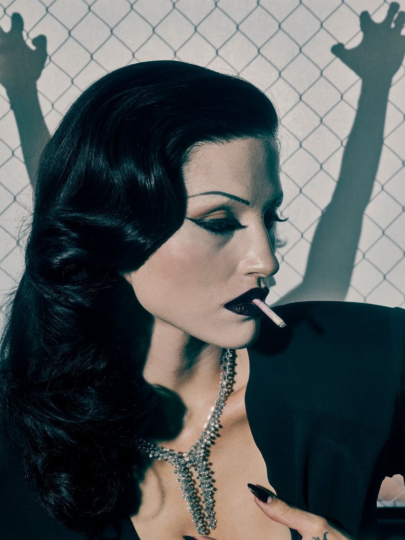 Side profile of a woman with dark hair, dark lipstick, and a necklace, smoking a cigarette against a wall with a fence shadow.