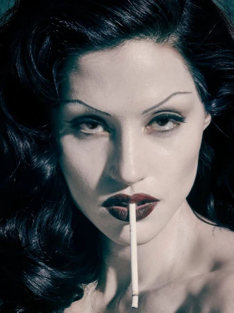 Portrait of a woman with dark hair and pale skin holding a cigarette. The image features dark, theatrical makeup and dramatic lighting.