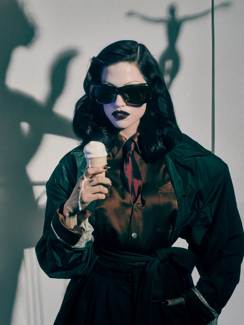 Elegant woman with dark hair and sunglasses poses in front of white wall, holding ice cream cone. Moody lighting and shadow figure.
