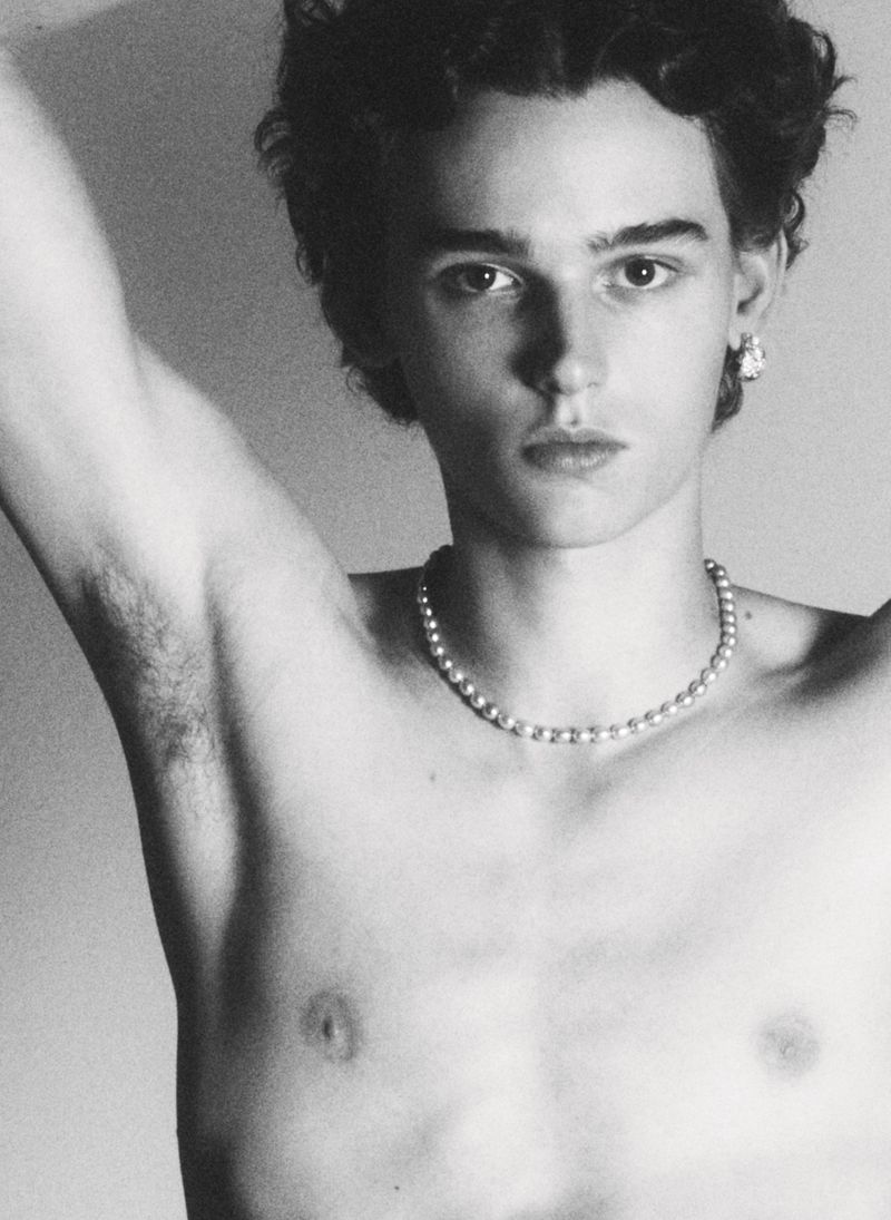 Monochrome portrait of a young adult male with curly dark hair wearing a pearl necklace and an earring with arm raised indoors.