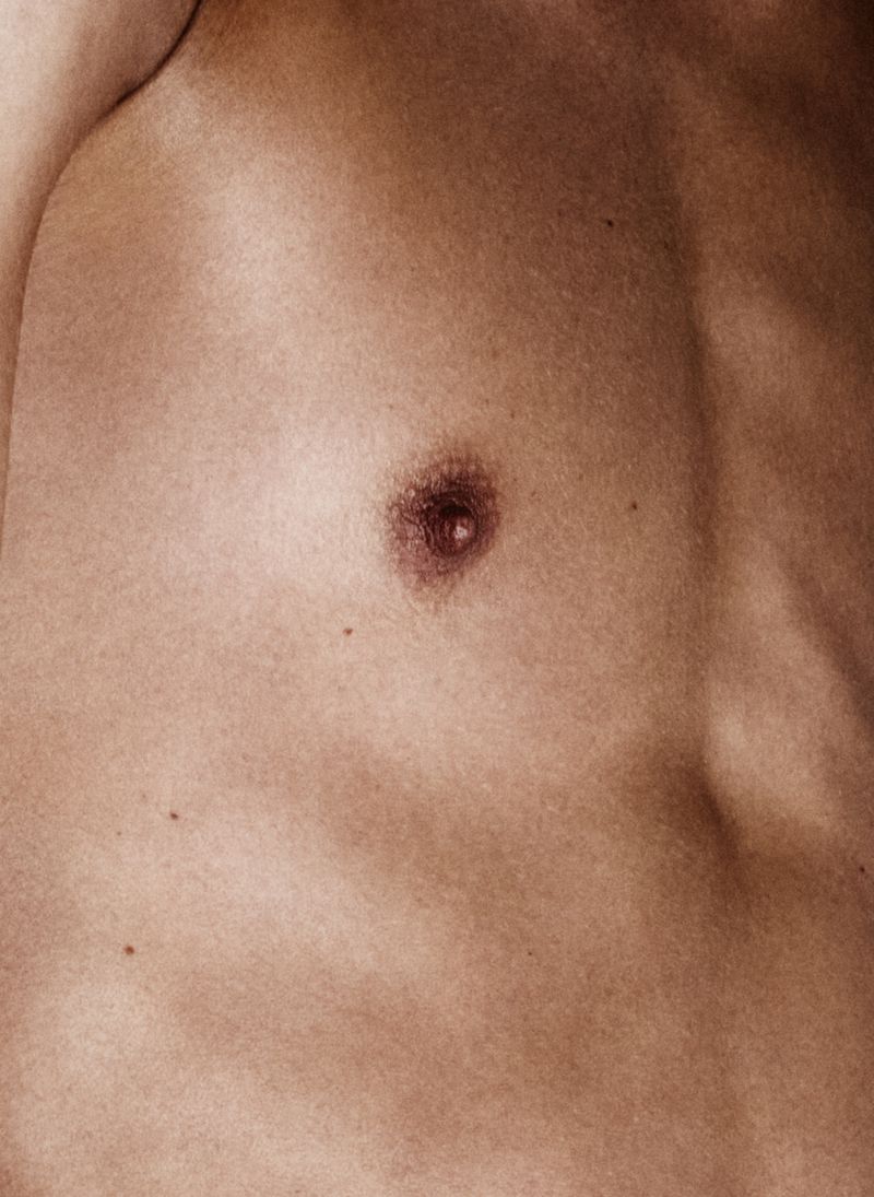 Close-up shot of the upper torso of a shirtless adult male, highlighting skin texture, muscle definition, and body details in a warm color palette.
