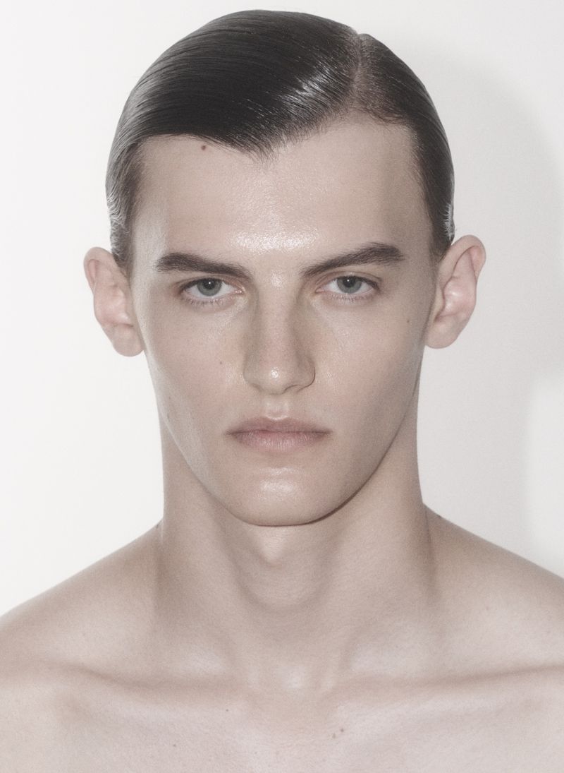 Close-up portrait of a young adult male with slicked-back dark hair and bare chest, standing in front of a plain white background.