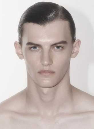 Close-up portrait of a young adult male with slicked-back dark hair and bare chest, standing in front of a plain white background.