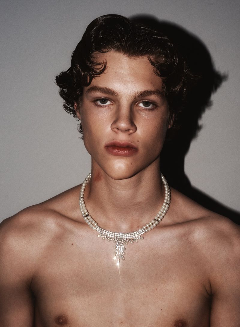 Shirtless male model wearing an ornate pearl and crystal choker necklace against a grey backdrop with direct flash shadow.