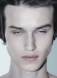Close-up portrait of a young male model with dark hair and pale skin against a neutral background.