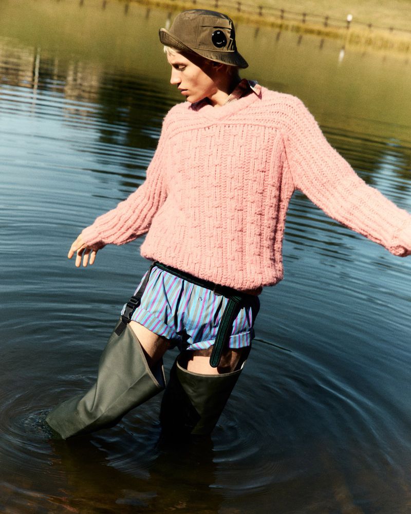 Man stands in lake wearing a pink sweater, striped shorts, olive fisherman hat, and tall rubber boots against an outdoor background.