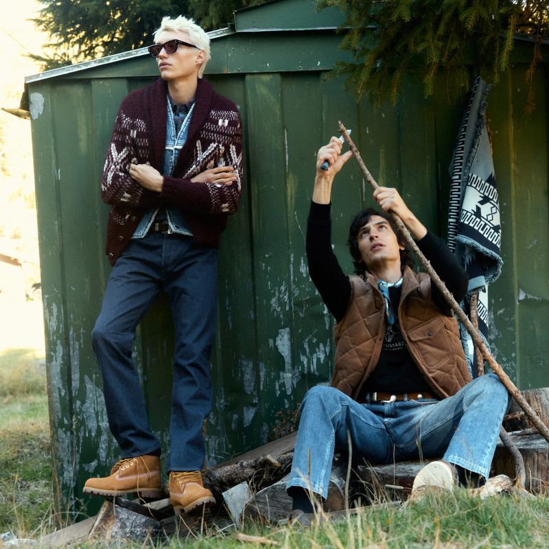 Two adult males pose outdoors near a green shed, one sharpening a stick, wearing casual fashion; fall colors and natural light.