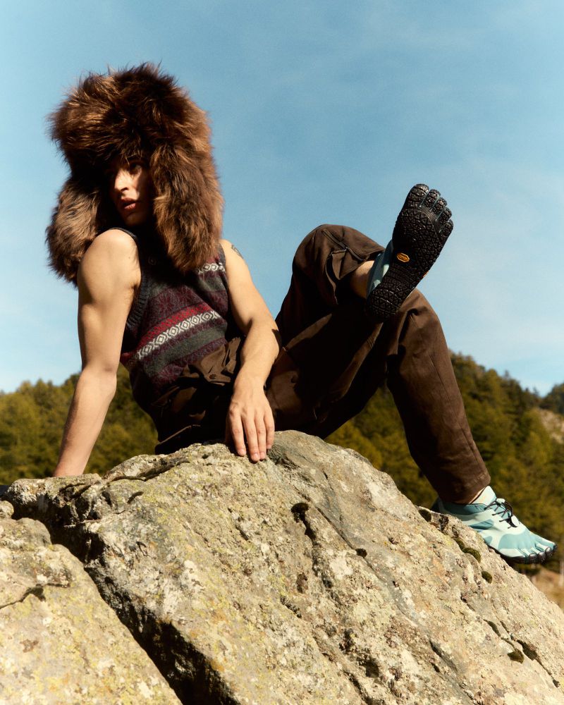 Person in a fur hat and patterned vest sits on a rock in the daytime, wearing toe shoes and brown pants; forested hills in the background.