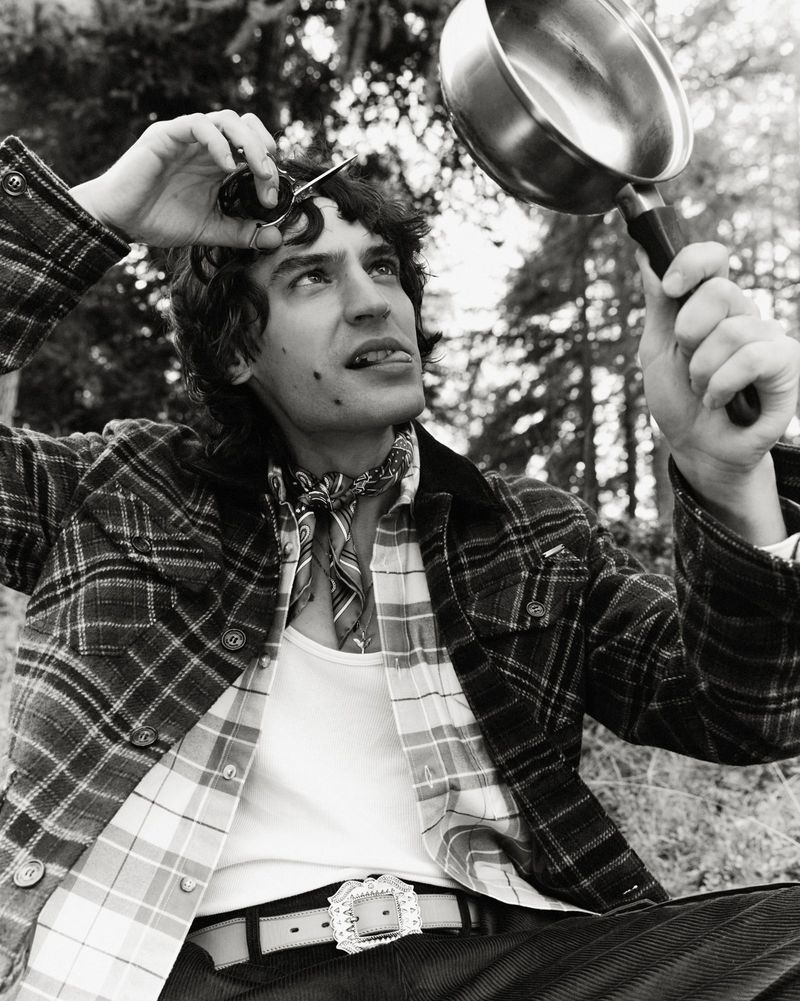 Man in a plaid jacket holds sunglasses and a frying pan outdoors with trees in background in a black and white, eye-level outdoor shot.