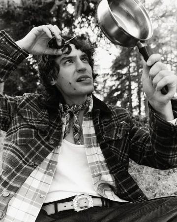 Man in a plaid jacket holds sunglasses and a frying pan outdoors with trees in background in a black and white, eye-level outdoor shot.