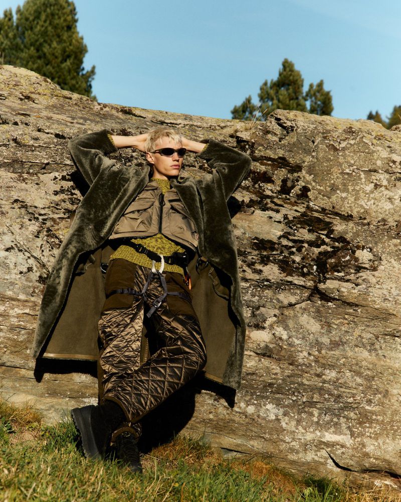 Adult male model lies against a rock wearing sunglasses, a long olive coat, vest, sweater, and quilted brown pants in outdoor setting.
