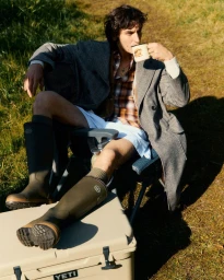 Man sitting on an outdoor chair outdoors wearing a grey coat, plaid shirt, shorts, and tall rubber boots while drinking from a mug