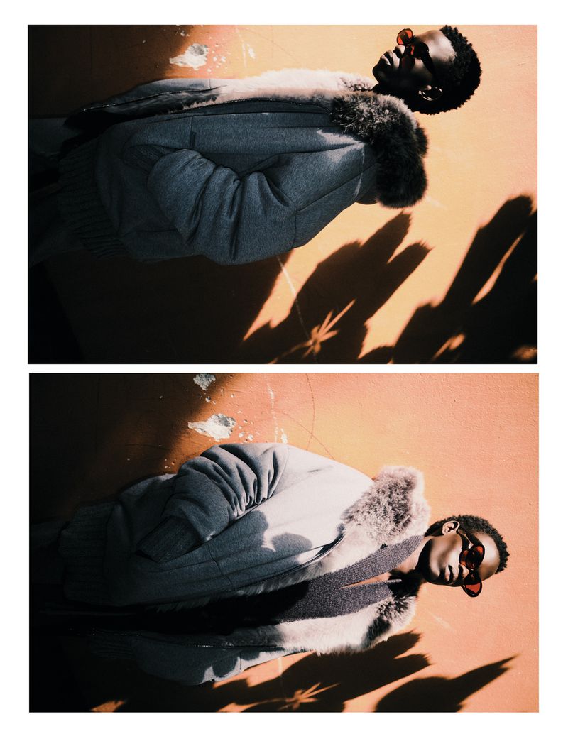 A person with dark skin and dark hair, is lying on a peach colored concrete surface with shadows across them, wearing a gray coat and red glasses.