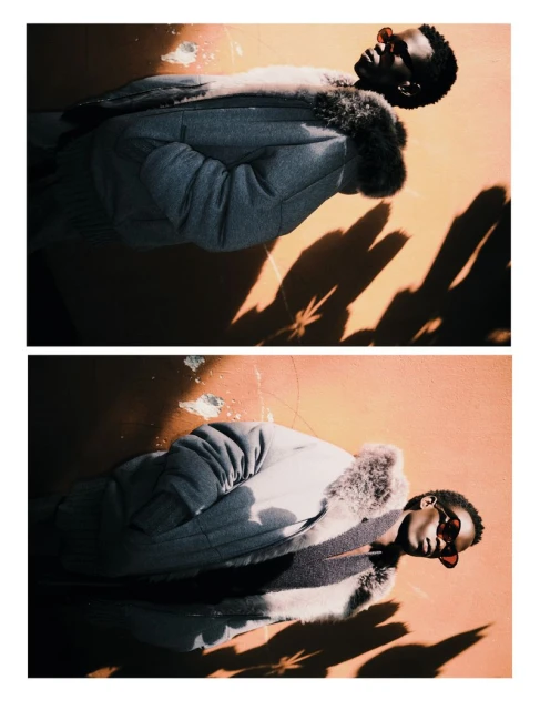 A person with dark skin and dark hair, is lying on a peach colored concrete surface with shadows across them, wearing a gray coat and red glasses.