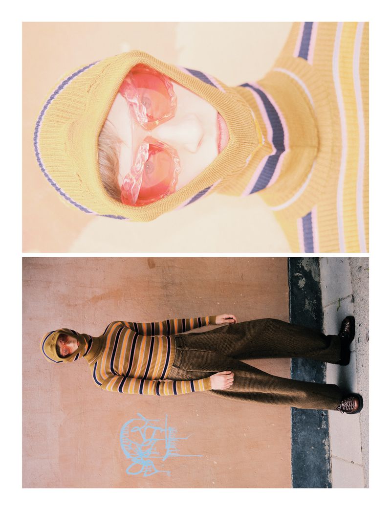 High-fashion editorial shot of a model wearing a striped turtleneck, brown trousers, and red sunglasses, lying on a concrete surface.
