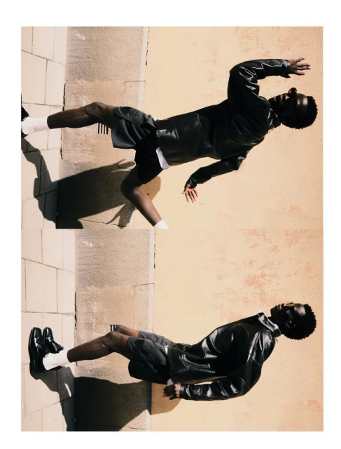 Fashion shot of an African-American male model in a black leather jacket, shorts, and white socks posing against a neutral wall outdoors.
