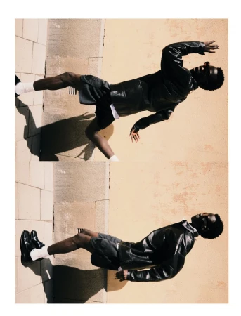 Fashion shot of an African-American male model in a black leather jacket, shorts, and white socks posing against a neutral wall outdoors.