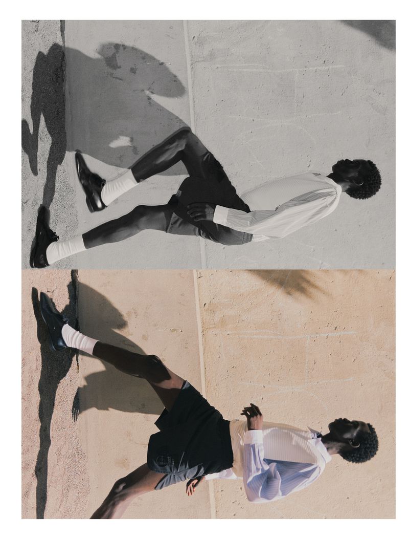 Fashion shot of an adult man lying on a cement surface wearing a white long-sleeved shirt, black shorts, white socks, and black loafers.