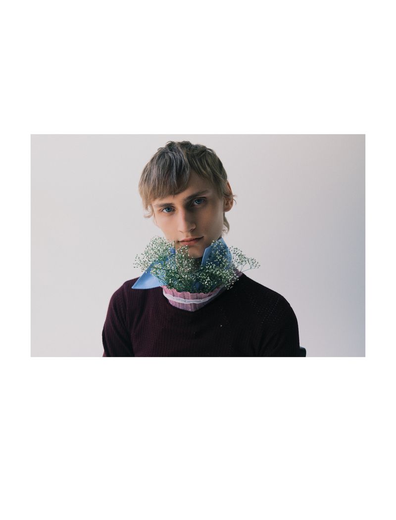 A young adult male with blond hair poses indoors wearing a maroon sweater and a light blue collared shirt with white baby's breath.