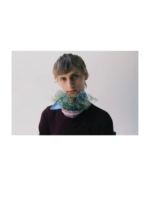 A young adult male with blond hair poses indoors wearing a maroon sweater and a light blue collared shirt with white baby's breath.