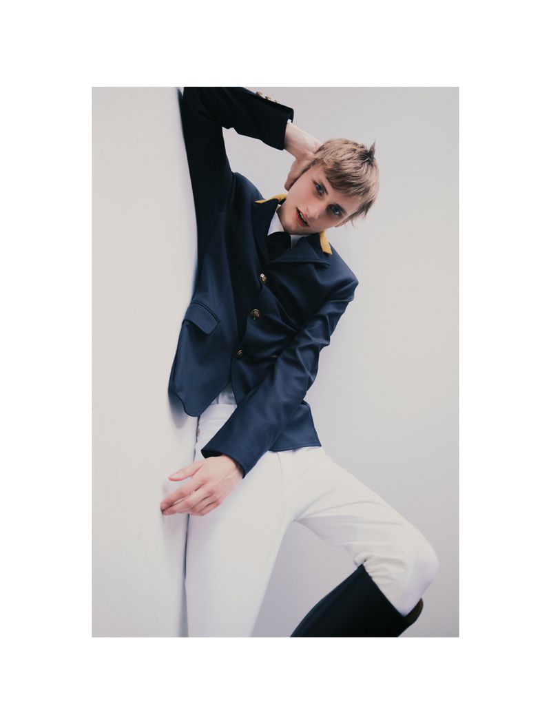 Young adult poses in a navy blazer with gold buttons and white trousers against a stark white wall in a fashion-style indoor photo.