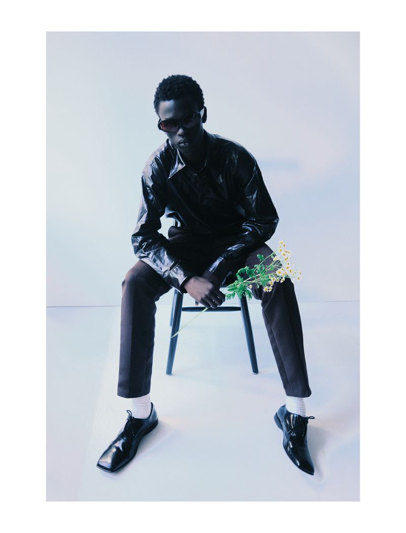 Dark-skinned male model in sunglasses and black jacket sits on chair holding flowers in an indoor setting with minimal background.