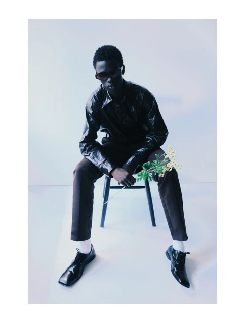 Dark-skinned male model in sunglasses and black jacket sits on chair holding flowers in an indoor setting with minimal background.