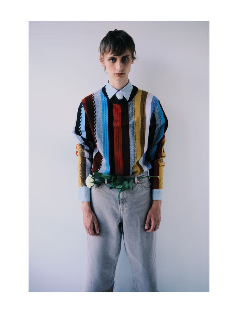 Man in a vertically striped colorful sweater stands against a white wall, accessorized with a white rose and light-wash jeans.