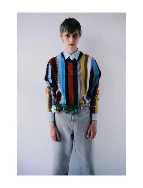Man in a vertically striped colorful sweater stands against a white wall, accessorized with a white rose and light-wash jeans.