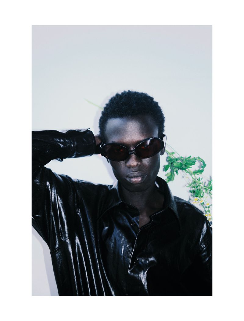 An adult male with dark skin wears sunglasses and a shiny black shirt. He poses with a white background and some greenery behind him.