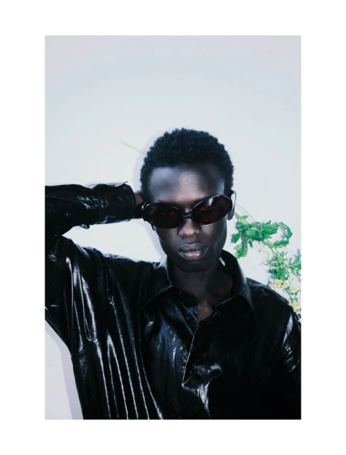 An adult male with dark skin wears sunglasses and a shiny black shirt. He poses with a white background and some greenery behind him.