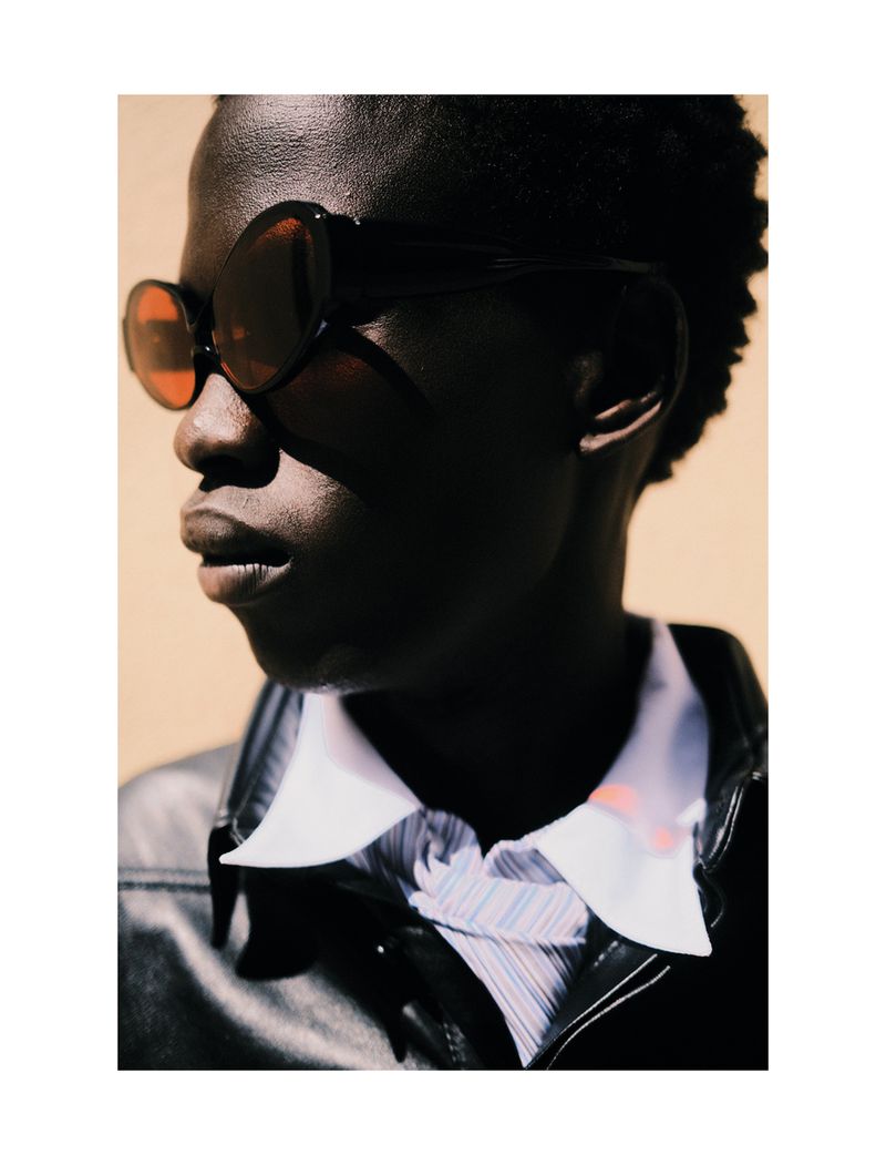 Close-up portrait of a Black man wearing dark oval sunglasses and a black leather jacket against a warm beige background.