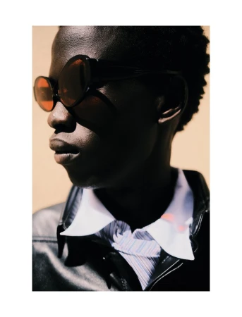 Close-up portrait of a Black man wearing dark oval sunglasses and a black leather jacket against a warm beige background.