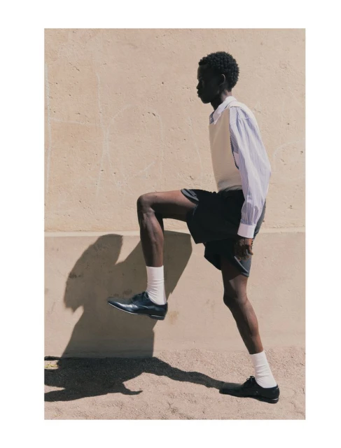 Dark-skinned adult male in fashionable clothes mid-stride outside on tan pavement near a tan wall on a sunny day with shadow detail.