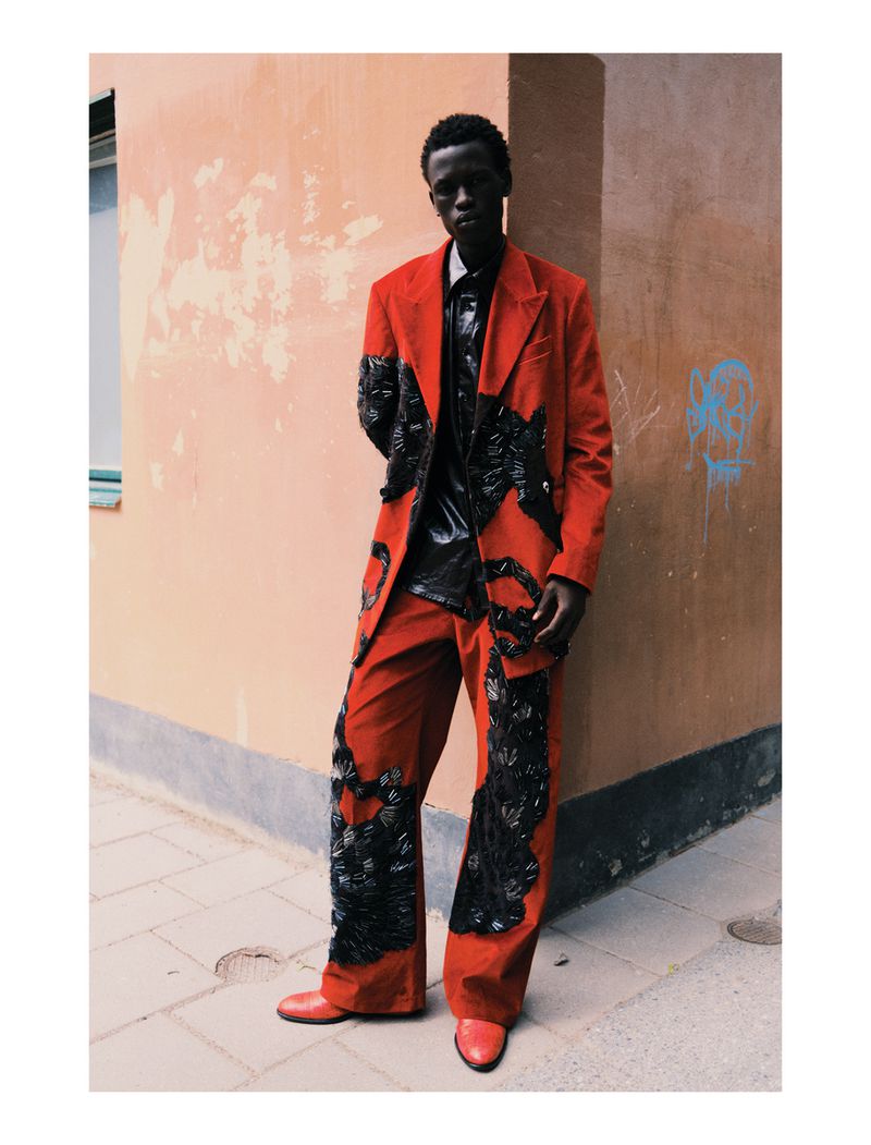 A dark-skinned adult male poses outdoors wearing an ornate red suit with black embellishments, a black shirt, and red shoes, leaning against a peach wall.